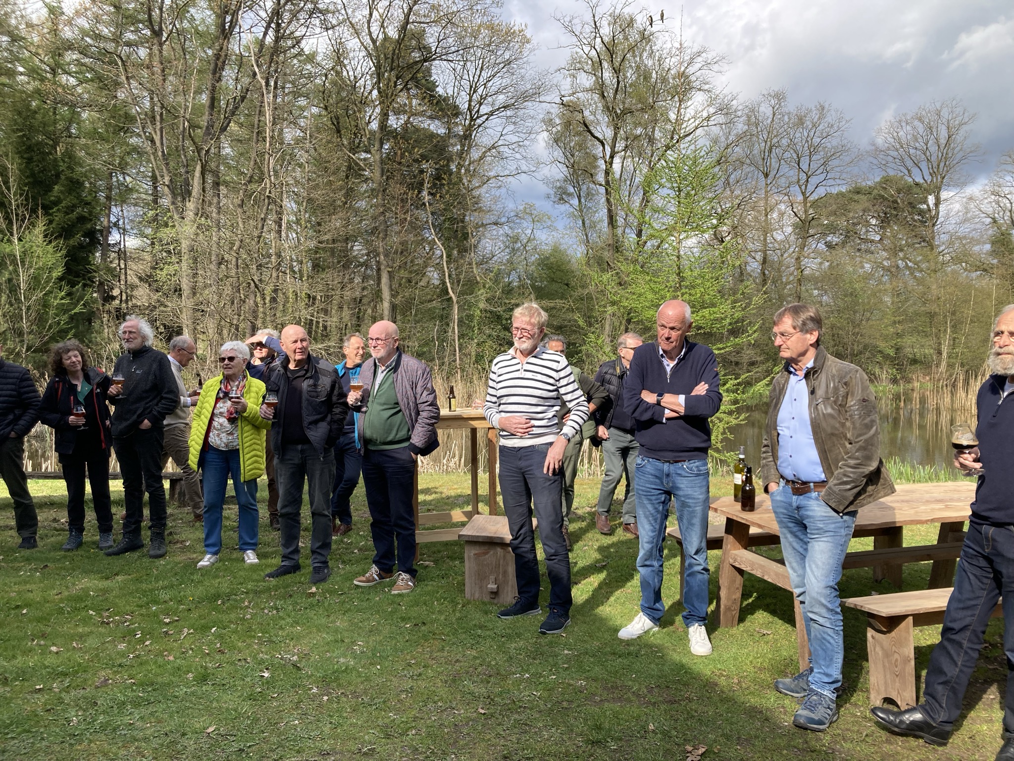 Een groep mensen die buiten is samengekomen, staand bij houten tafels met op de achtergrond een waterpartij.