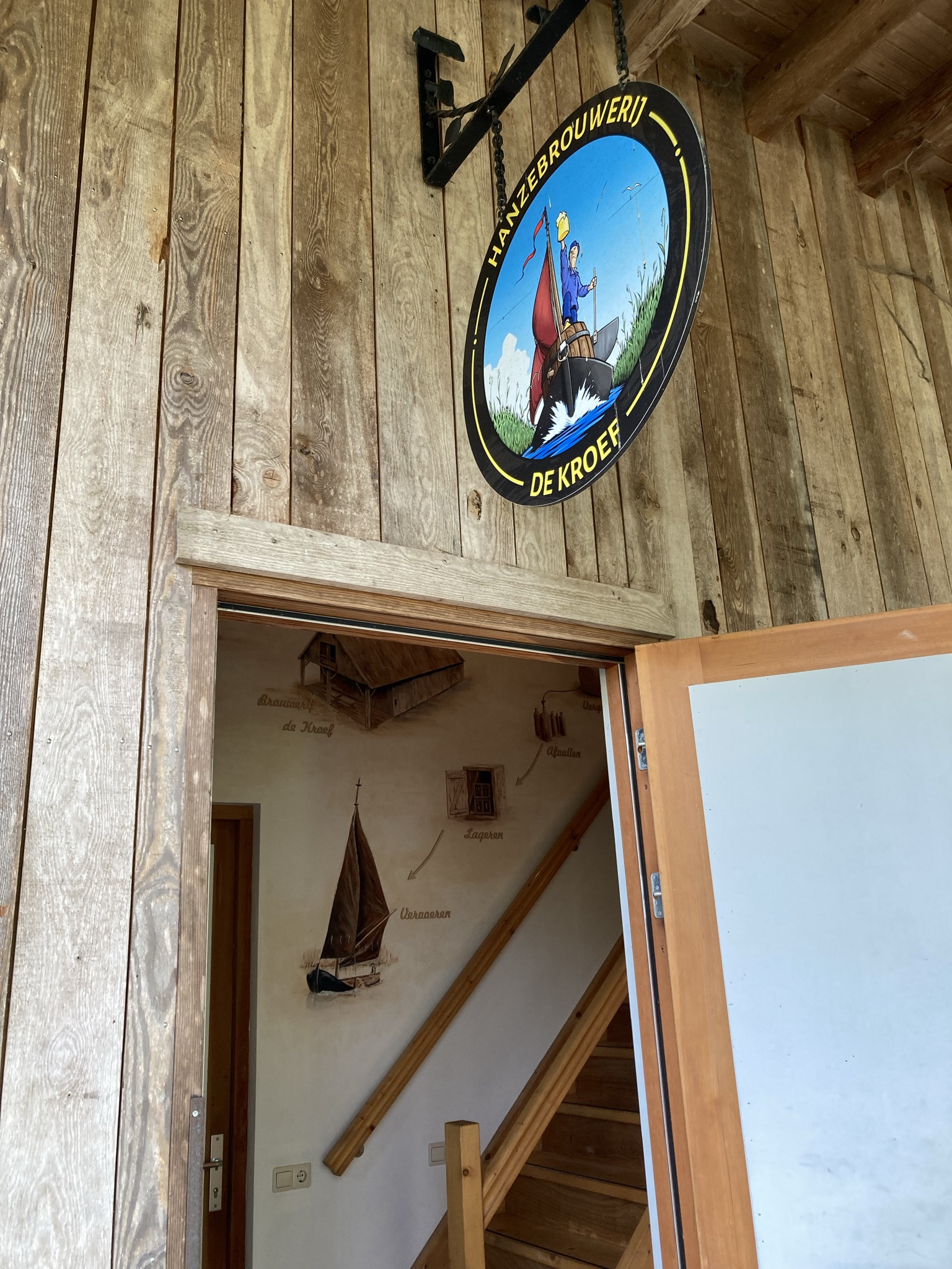 Een ingang van een gebouw met een rond bord voor 'Hanzebrouwerij de Kroef' en een glimp van de muurschildering binnen.